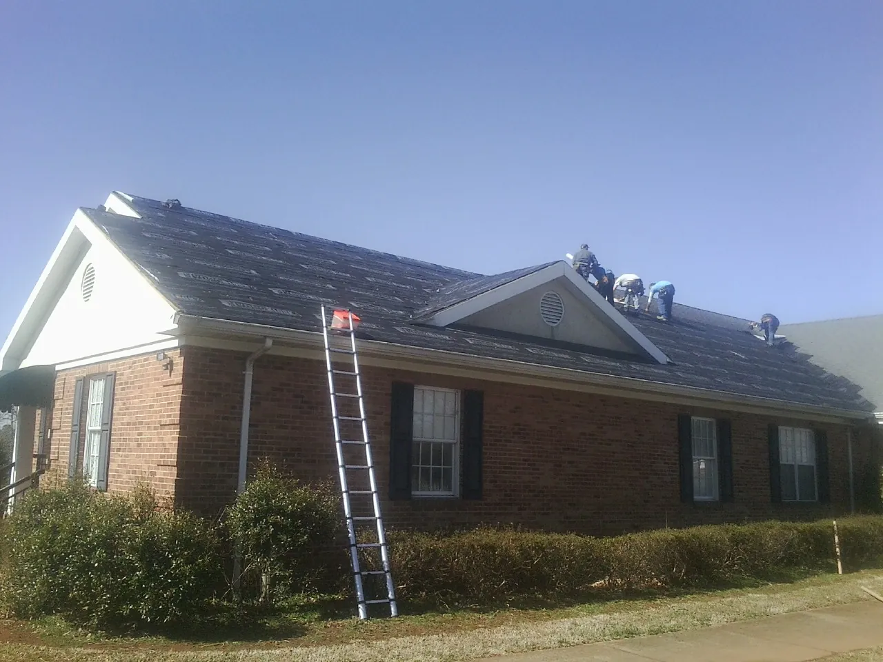 Crew tearing shingles off a brick ranch roof with a ladder set — Emergency Roof Repair work by our crew in Chapman
