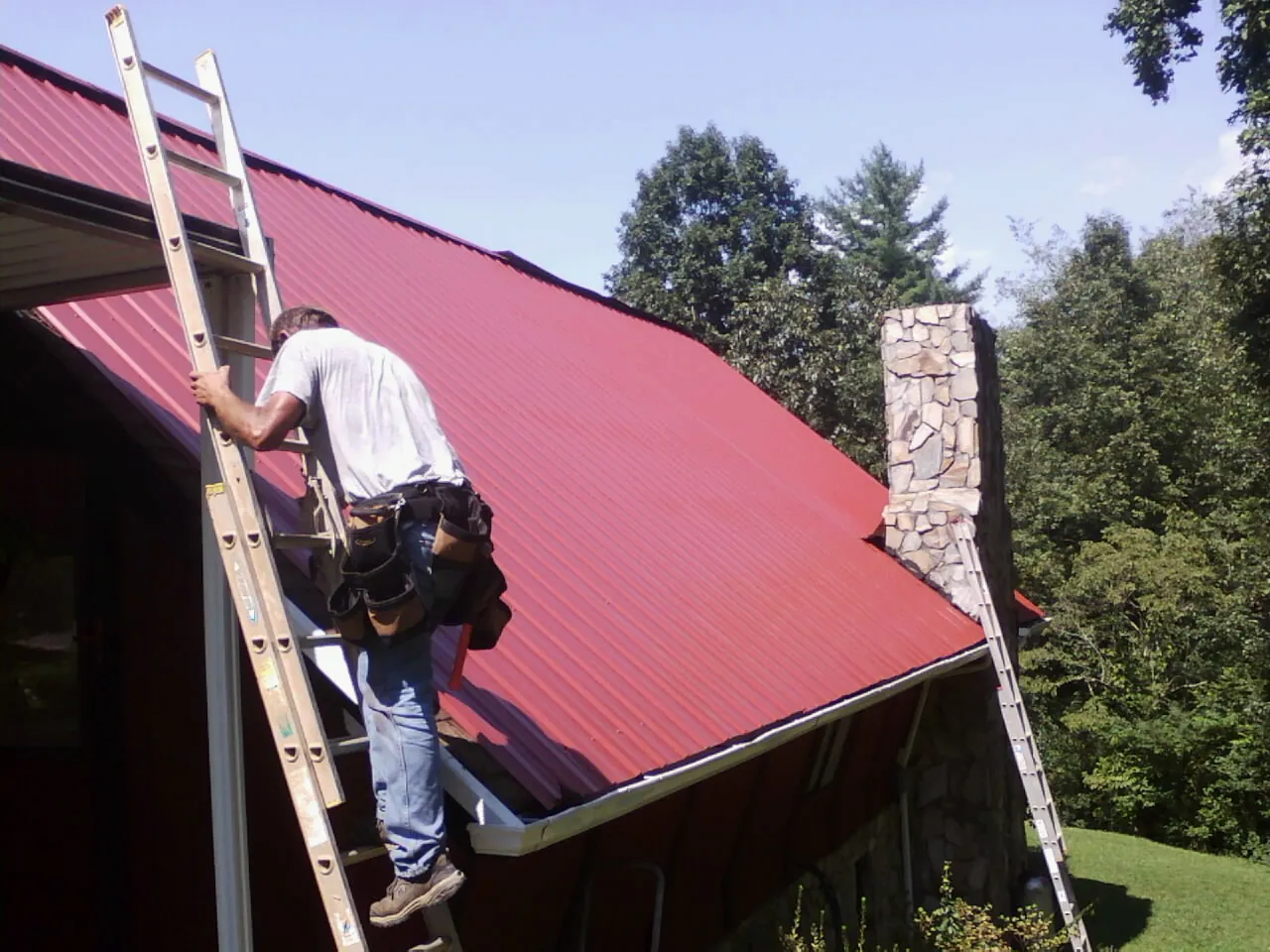 Roofer on a ladder beside a red metal roof and stone chimney — Soffit & Fascia Repair by Roofing Brothers in Chapman